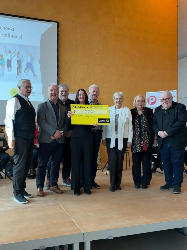 Eine Gruppe von Menschen steht auf einer Bühne, einer hält einen gelben Scheck über 10.000 Euro. Sie sind formell gekleidet, wahrscheinlich für eine Zeremonie. Hinter ihnen zeigt eine Leinwand Bilder von tanzenden Menschen.