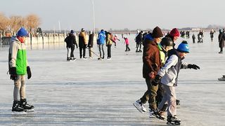 A group of people ice skating on a frozen lake, some wearing helmets and holding hockey sticks, with others standing around watching.