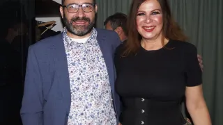 Ein Mann und eine Frau stehen eng beieinander und lächeln für ein Foto. Der Mann trägt eine Brille und ein Blumenhemd, während die Frau lange Haare und ein schwarzes Oberteil mit einem Blumenrock hat. Hinter ihnen ist ein Vorhang und der Kopf eines Mannes zu sehen.