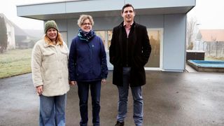 Three people stand outside in front of a building. Two women and one man, all smiling. The women wear jackets and the man wears a black coat.