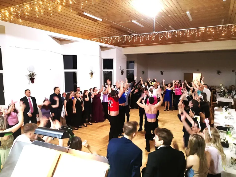 Eine große Gruppe von Menschen tanzt in einem Raum mit Holzböden, einige in Kostümen. Andere schauen zu und jubeln. Der Raum hat weiße Wände, eine Holzdecke mit Lichtern und Fenster.