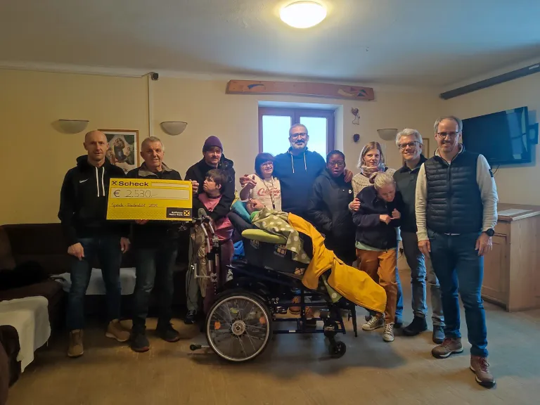 Eine Gruppe von Menschen posiert mit einer Person im Rollstuhl und einem gelben Scheck über 2530 Euro. Sie befinden sich in einem Raum mit einer Holzwand und einem Fenster.