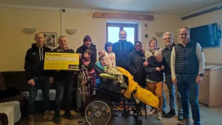 A group of people pose with a person in a wheelchair and a yellow check for 2530 Euros. They are in a room with a wooden wall and a window.