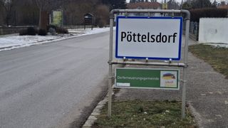 A street sign for Pöttelsdorf is on the side of the road. The road is empty with some grass on the side.
