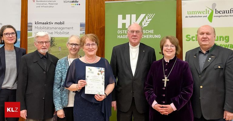 Eine Gruppe von Menschen steht zusammen und hält eine Auszeichnung, ein Mann in einem Anzug steht zwischen zwei Frauen, eine von ihnen hält ein Zertifikat. Hinter ihnen ist ein Banner mit grünem Text und Symbolen.