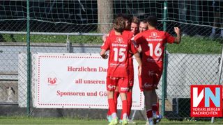 Soccer players in red uniforms celebrate a goal in front of a fence with a sponsor banner reading 'Herzlichen Dank all unseren Sponsoren und Gönnern.' Players wear numbers 15, 16, and 17.