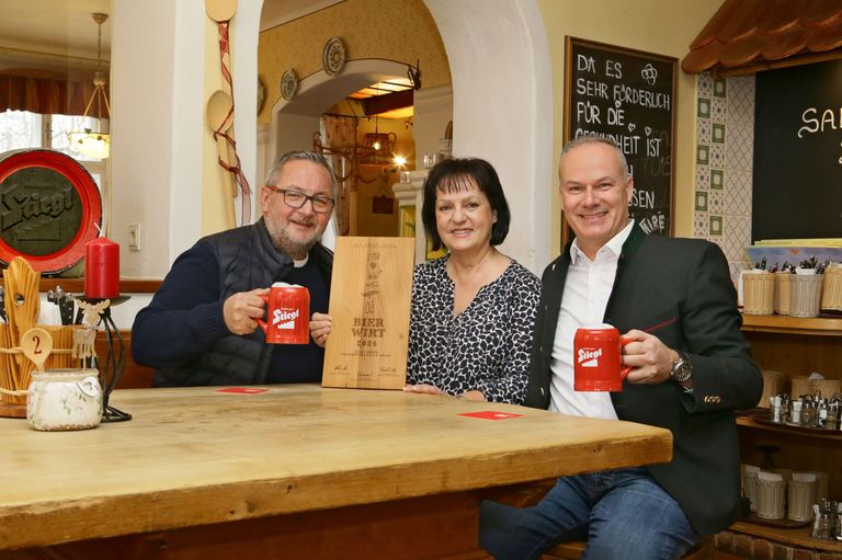 Drei Personen sitzen an einem Tisch, zwei Männer und eine Frau, halten rote Tassen. Eine Holztafel mit 'BIER WÜRT 2026' steht auf dem Tisch. Der Hintergrund besteht aus einer gelben Wand mit Dekorationen und einer Tafel.