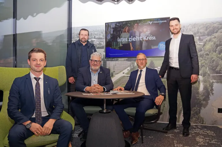 Fünf Männer in Anzügen posieren für ein Foto in einem Büro. Ein Mann steht und lächelt, während die anderen um einen Tisch herumsitzen. Ein Bildschirm hinter ihnen zeigt eine Werbung für Volkswagen Bank.