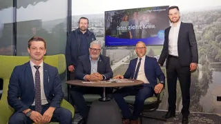 Five men in business suits pose for a photo in an office. One man stands, smiling, while the others sit around a table. A screen behind them displays a Volkswagen Bank advertisement.
