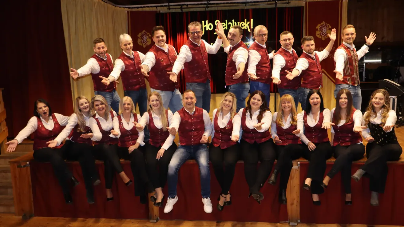 Eine Gruppe von Erwachsenen, sowohl Männer als auch Frauen, gekleidet in passenden roten Westen und Jeans, posiert für ein Foto auf einer Bühne mit rotem Vorhang im Hintergrund.