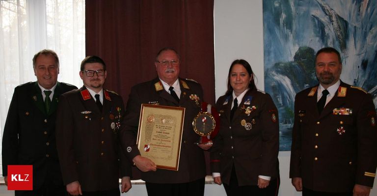 Vier Menschen in Militäruniformen stehen vor einer Wand. Der Mann in der Mitte hält eine gerahmte Auszeichnung und eine Trophäe. Die beiden Frauen stehen auf beiden Seiten von ihm. Dahinter ist ein Gemälde eines Wasserfalls.