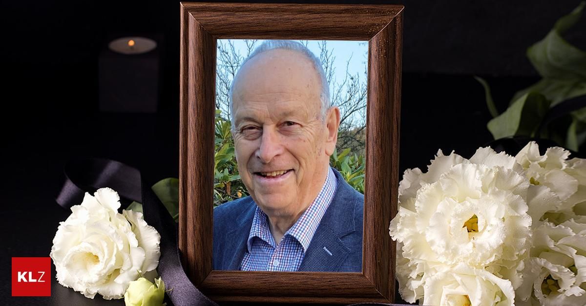 A photo frame with a smiling elderly man is placed on a black background. The man is wearing a blue suit and a white checkered shirt. On the left is a white flower, and on the right is a black ribbon.