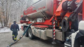 A firefighter is attaching a hose to a red and silver truck labeled 'FEURWEHR' with a capacity of 12,000 liters. The scene is set in a snowy, wooded area.