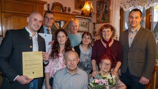 An elderly couple stands surrounded by family members in a living room, holding a certificate and flowers. The room is decorated with framed pictures, a clock, and a lamp.
