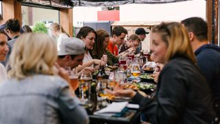 A group of people are seated at a long table, possibly at a restaurant, and they are enjoying their meals and drinks. They are smiling and talking to each other.