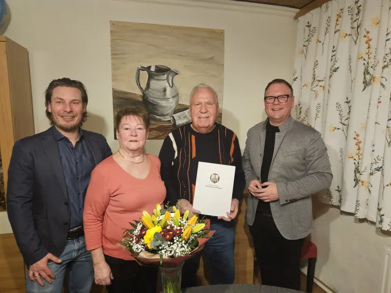 Vier Menschen stehen lächelnd zusammen im Innenbereich. Zwei Männer halten ein Zertifikat und einen Blumenstrauß. Ein Gemälde einer Kanne befindet sich hinter ihnen.