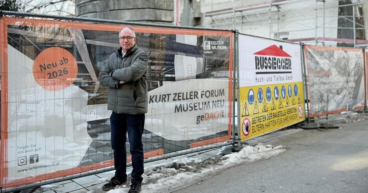 A man stands by a fence with banners for Kelten Museum Hallein and Kurt Zeller Forum. He is wearing a jacket and glasses.