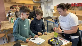 Three young boys are working on a project at a wooden table. One holds scissors, another holds a box, and a girl watches. They are in a workshop with shelves, a sink, and other people in the background.