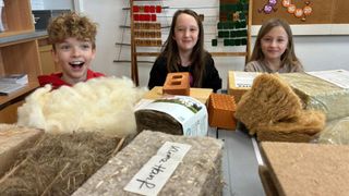 Three children in a classroom, one smiling and two others looking at woolen and wooden materials on a table.