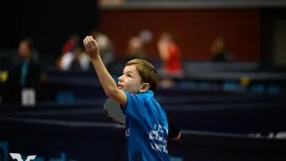 A boy plays table tennis, aiming to hit a ball in mid-air. He wears a blue shirt with white text. In the background, blurred spectators are watching.