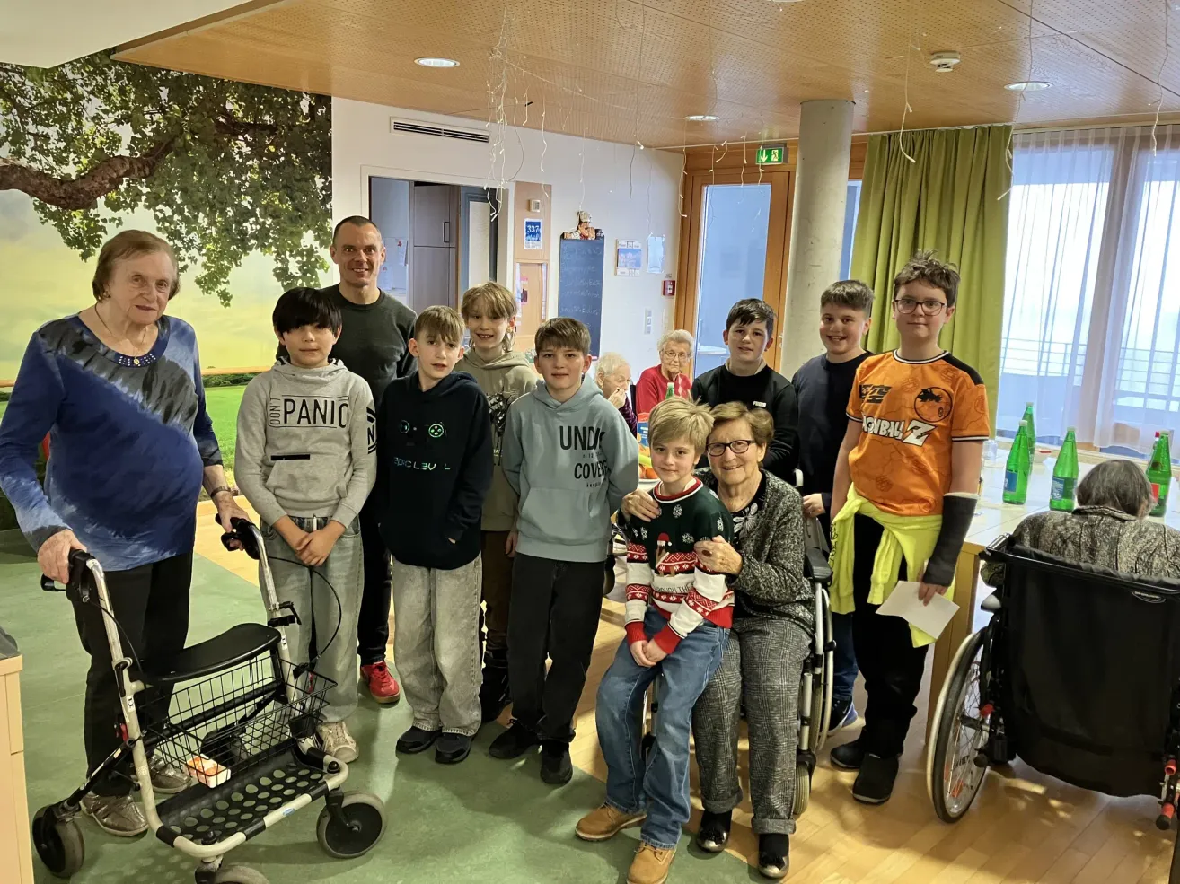 Eine Gruppe von Kindern und älteren Menschen versammelt sich in einem Raum, lächelt und posiert für ein Foto. Eine Frau im Rollstuhl hält einen Jungen auf dem Schoß. Eine Person hält einen Gehwagen.