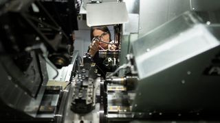 A woman wearing glasses and a black shirt is working on a machine in a factory. The machine has many mechanical parts, and she is holding a tool in her hand.