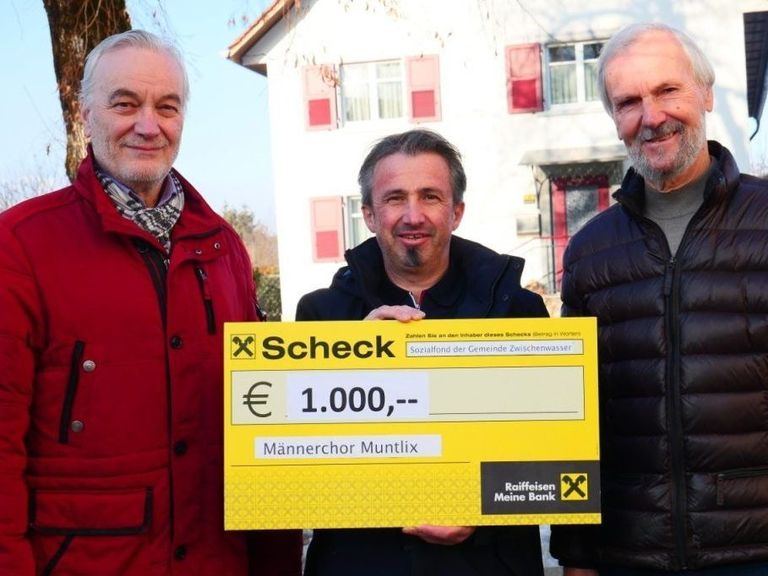 Drei Männer stehen vor einem Haus und lächeln. Der Mann in der Mitte hält einen großen Scheck über 1.000 Euro vom Sozialfonds der Gemeinde Zwischenwasser.