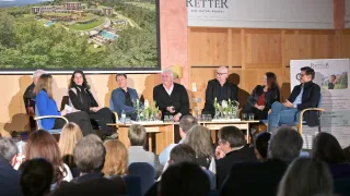 A panel of four people sits in front of a large screen displaying a resort. Glasses and papers are on the tables before them. The audience sits in front of them.