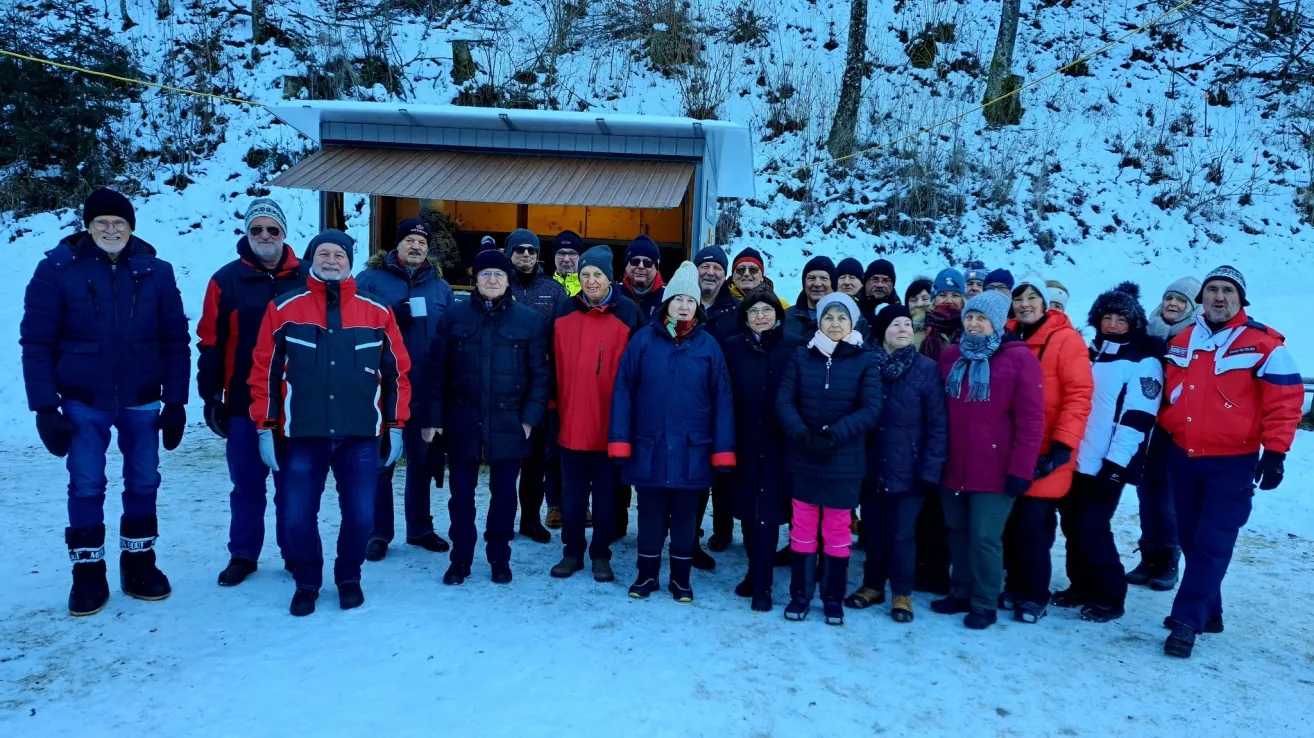 A group of people dressed in winter gear, including hats and gloves, are standing in the snow, smiling for a photo in front of a small building.