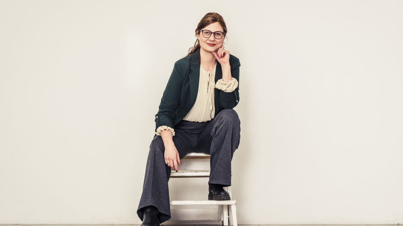 A woman wearing glasses, a black blazer, and gray pants is sitting on a white chair, smiling and posing for a photo.