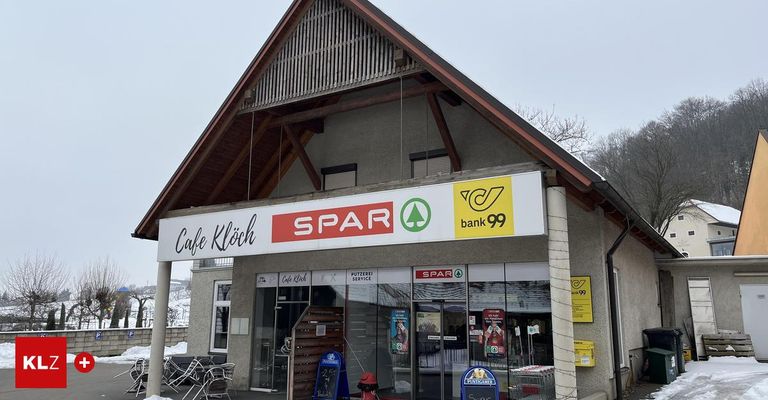 Ein Gebäude mit einem dreieckigen Dach hat ein Schild mit der Aufschrift SPAR und Cafe Kloch. Es hat Glastüren und Fenster mit Bannern. Draußen sind Stühle, ein Feuerlöscher und eine gelbe Box. Bäume stehen dahinter.