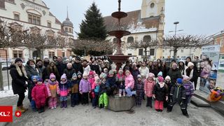 A group of children and adults pose for a photo in front of a fountain in a town square, surrounded by buildings and trees.