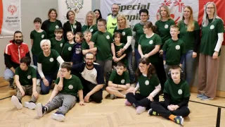 A group of adults and children in green shirts pose for a photo in a room with a wooden floor and a banner behind them.
