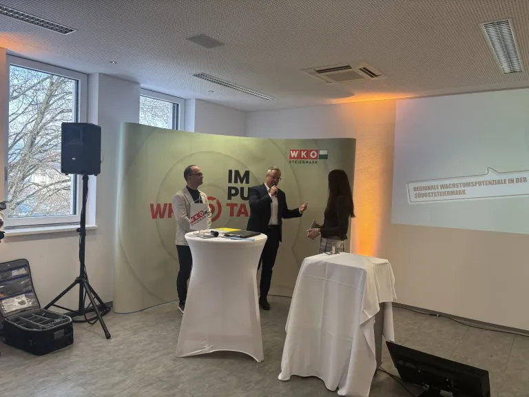 Three people stand in front of a banner with the text 'Im Pu Ta'. A man speaks into a microphone. Another man stands behind a desk with papers and a book. A woman stands next to him.