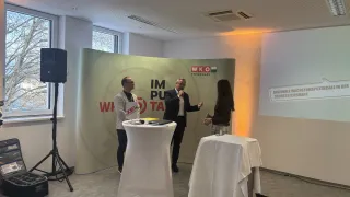 Three people stand in front of a banner with the text 'Im Pu Ta'. A man speaks into a microphone. Another man stands behind a desk with papers and a book. A woman stands next to him.