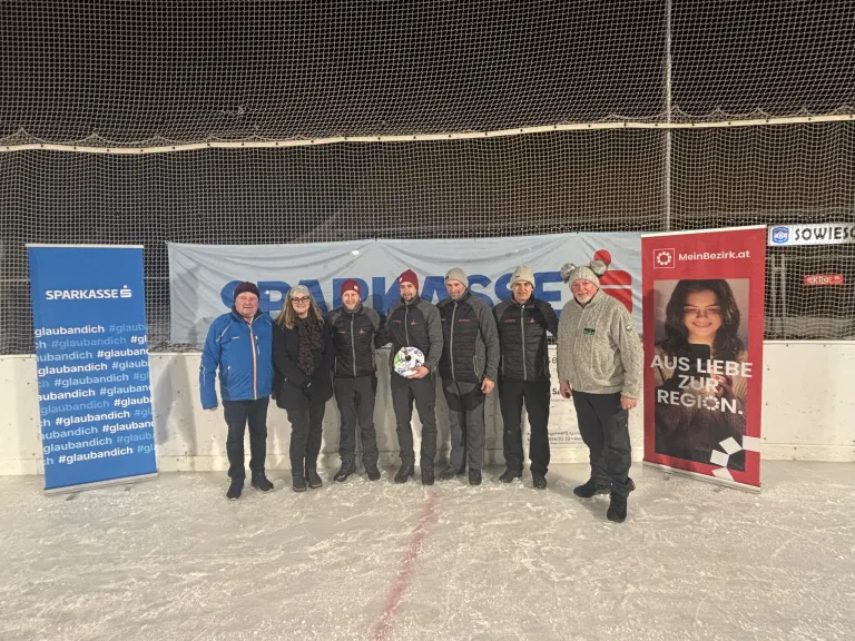 Eine Gruppe von Männern und Frauen steht auf einer Eisbahn und posiert für ein Foto. Sie tragen alle Winterkleidung. Ein Banner und ein Plakat stehen hinter ihnen.