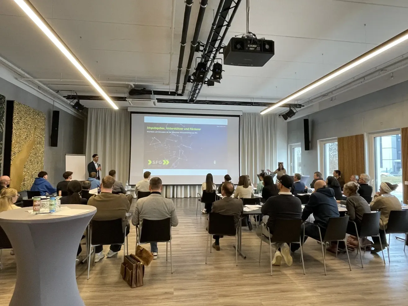 A group of people are sitting in a conference room, with a projector screen showing an image and text. A man is standing at a podium in front of them.
