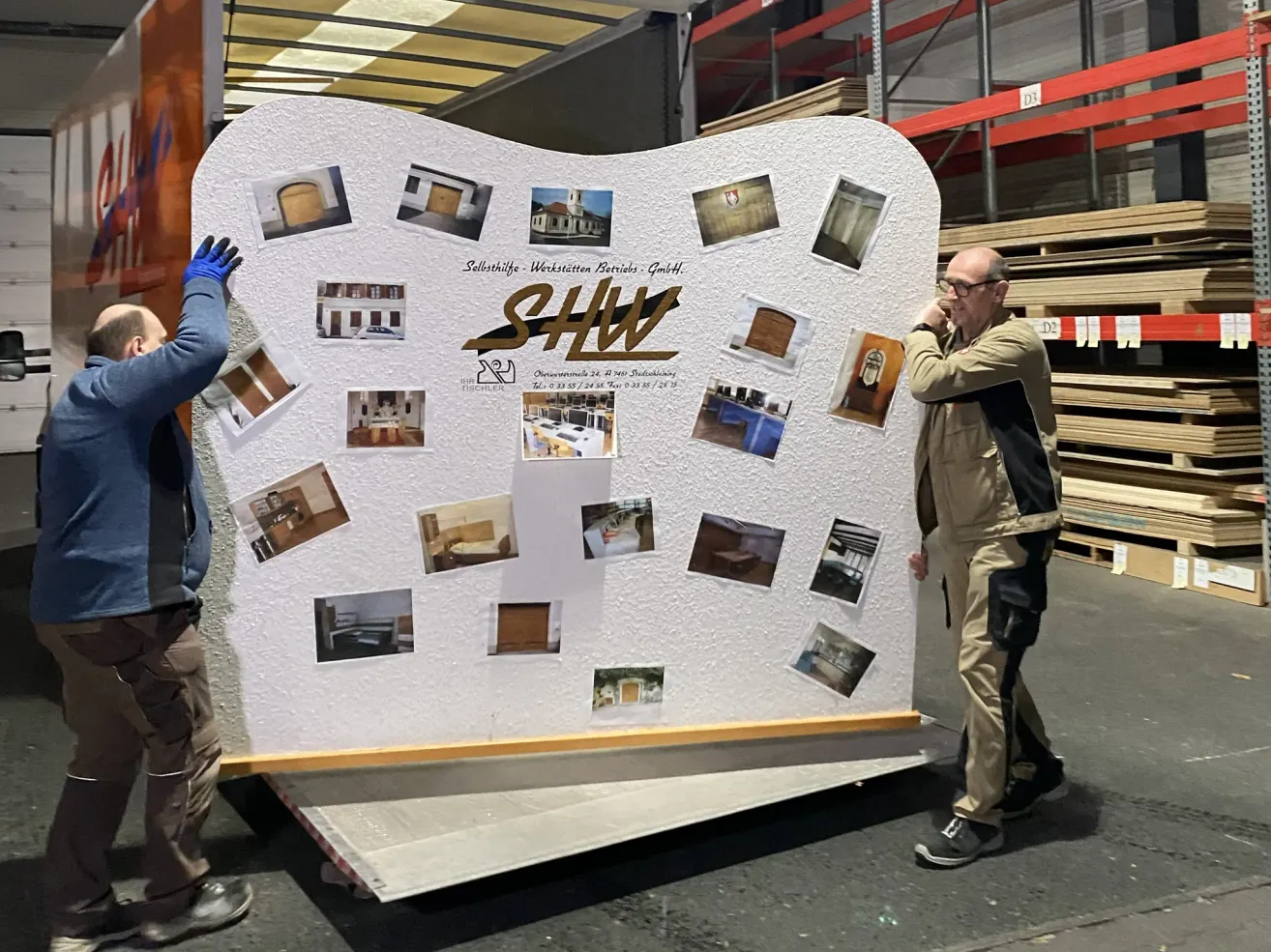Two workers are unloading a large white wall with various photographs attached to it. The wall features the logo SHWL. Behind the wall, there are wooden pallets stacked.