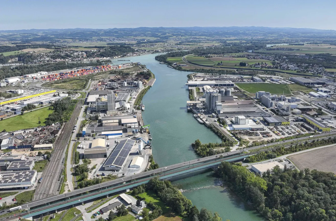 An aerial view of an industrial area by a river, with several factories, a bridge, and a train track. Trees and mountains are visible in the distance.