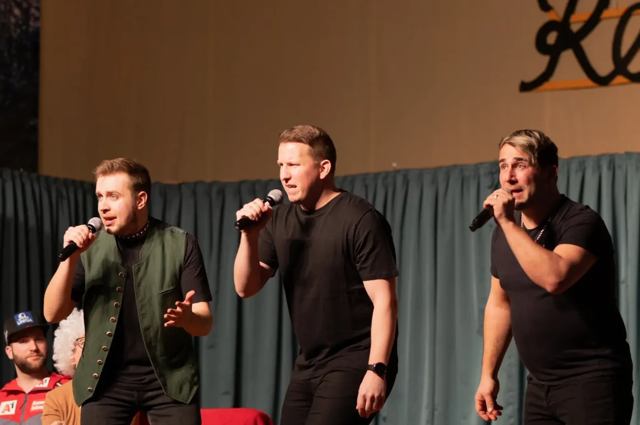 Three men are performing on stage. The man on the left wears a green vest and speaks to the audience. The center man holds a microphone and sings. The right man, wearing a black shirt, also sings.