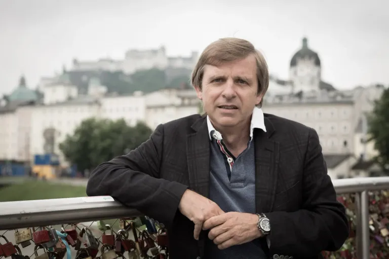 A man leans on a metal railing with numerous padlocks, wearing a black jacket and a watch, with a historical building in the background.
