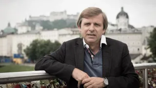 A man leans on a metal railing with numerous padlocks, wearing a black jacket and a watch, with a historical building in the background.