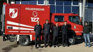 Four men stand in front of a red fire truck with the number 122. They wear black jackets and pants. Behind them is a building with glass windows.