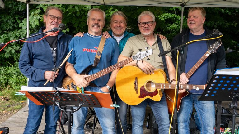 Fünf Männer posieren für ein Foto unter einem Baldachin. Sie lächeln und halten Gitarren. Einer von ihnen spielt einen Bassgitarre.
