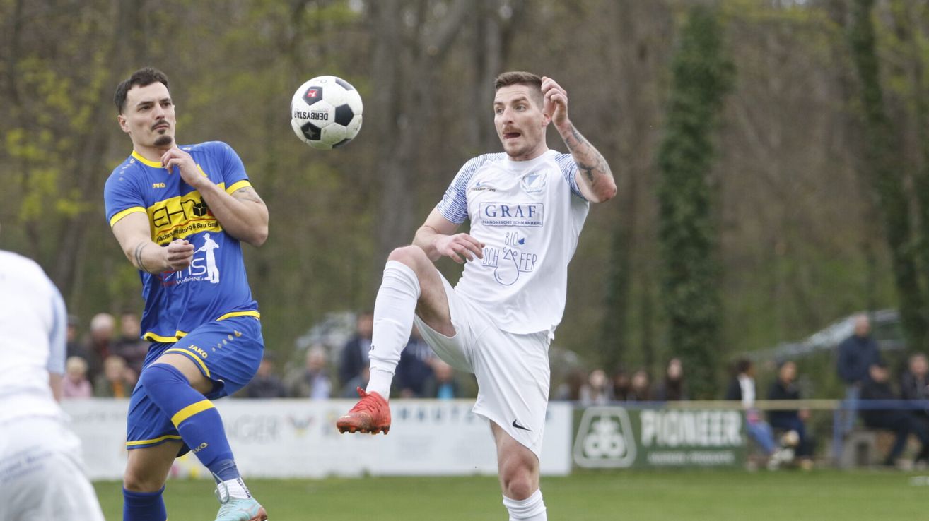 Ein Fußballspieler tritt den Ball, sein Teamkollege verteidigt und Zuschauer beobachten von hinten.