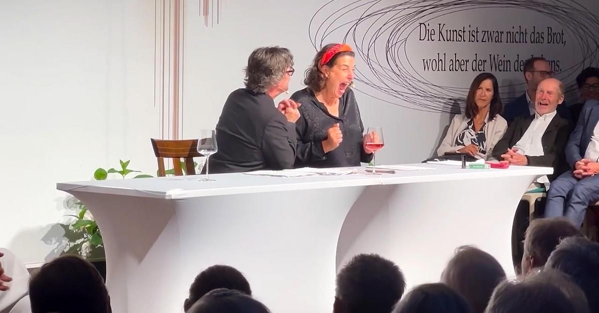 Three individuals seated at a table, two are engaged in conversation while the third listens. One holds a wine glass. Behind them is a wall with text. The setting appears formal.