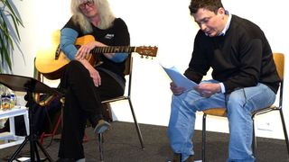 A woman with glasses and a guitar sits on a chair, while a man with a paper reads in front of her.