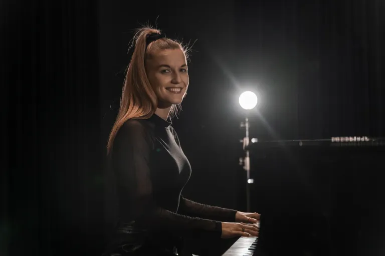 Eine Frau mit langen blonden Haaren spielt lächelnd ein Klavier. Sie trägt ein schwarzes Oberteil und einen Ring. Das Klavier ist schwarz, und ein helles Licht scheint auf sie. Hinter ihr hängt ein Vorhang an der Wand.