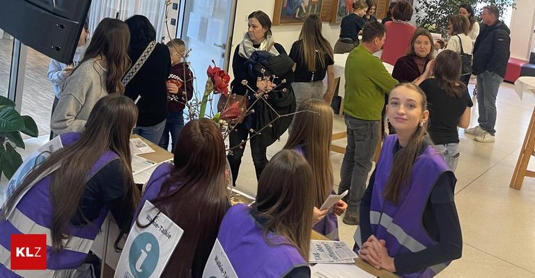 Eine Gruppe von Menschen in lila Westen versammelt sich in einem Raum, möglicherweise für eine Besprechung. Eine Frau mit einem Baby steht in der Mitte. Papiere und eine Blumenarrangement sind auf dem Schreibtisch.