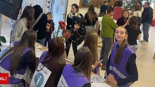 A group of people in purple vests are gathered in a room, possibly for a meeting. A woman holding a baby stands in the center. Papers and a flower arrangement are on the desk.